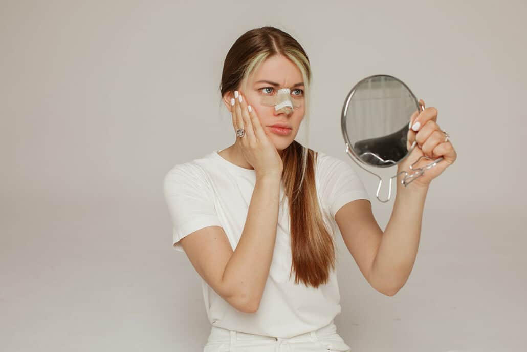 Beautiful female rhinoplasty patient looking herself in the mirror waiting for a secondary procedure.