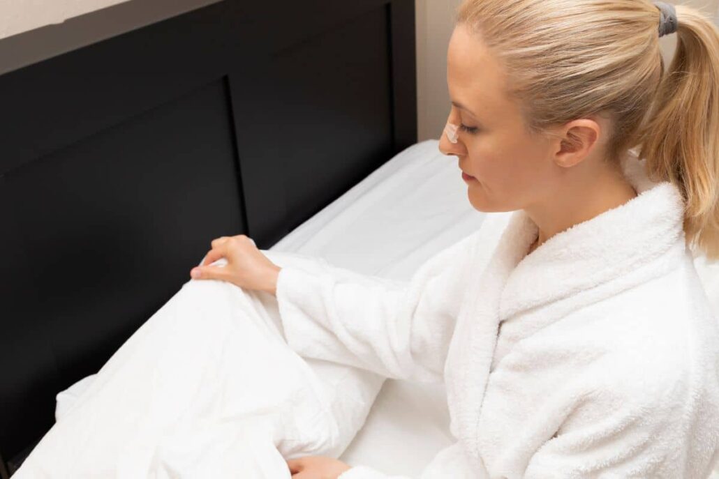 Woman with nasal bandage preparing bed with pillows to sleep elevated after rhinoplasty for comfort and healing.